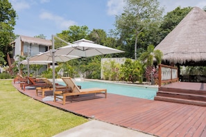 Un espace aménagé au bord de la piscine, avec des chaises longues en bois et des parasols, entouré d’une végétation luxuriante et agrémenté d’une structure à toit de chaume.