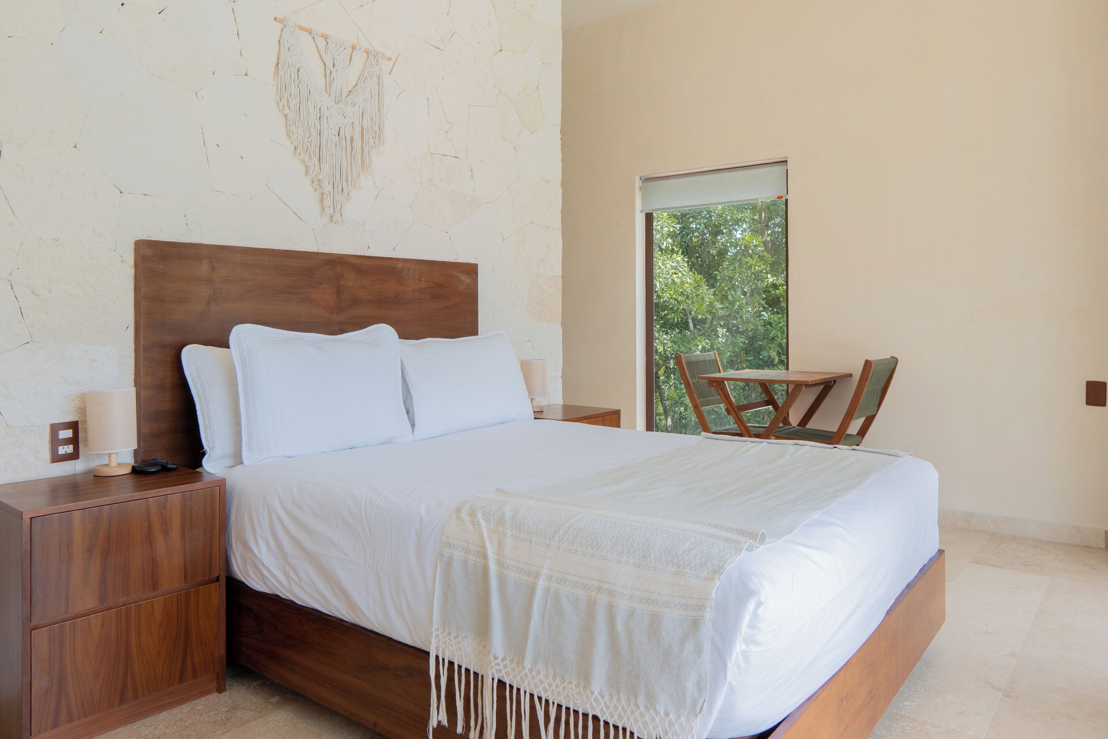 A bedroom with a wooden bed, a nightstand, a chair, and a window with a view of greenery.