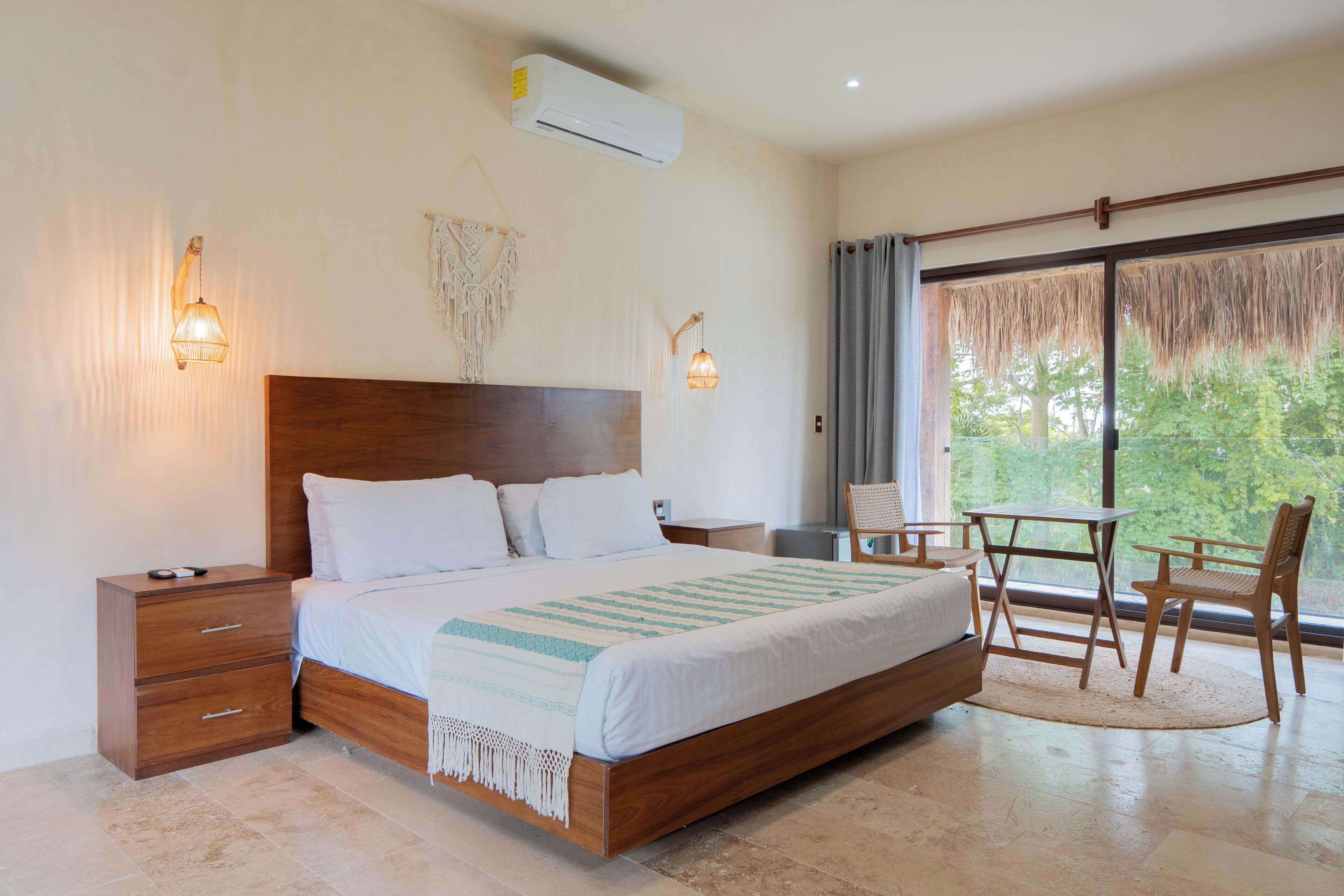 Un dormitorio con cama de madera, mesitas de noche, un escritorio, una silla y un amplio ventanal con vista a zonas verdes.