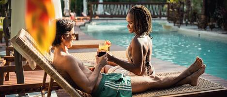 A couple relaxing by a poolside lounge chair with a view of the pool and a thatched-roof structure in the background.