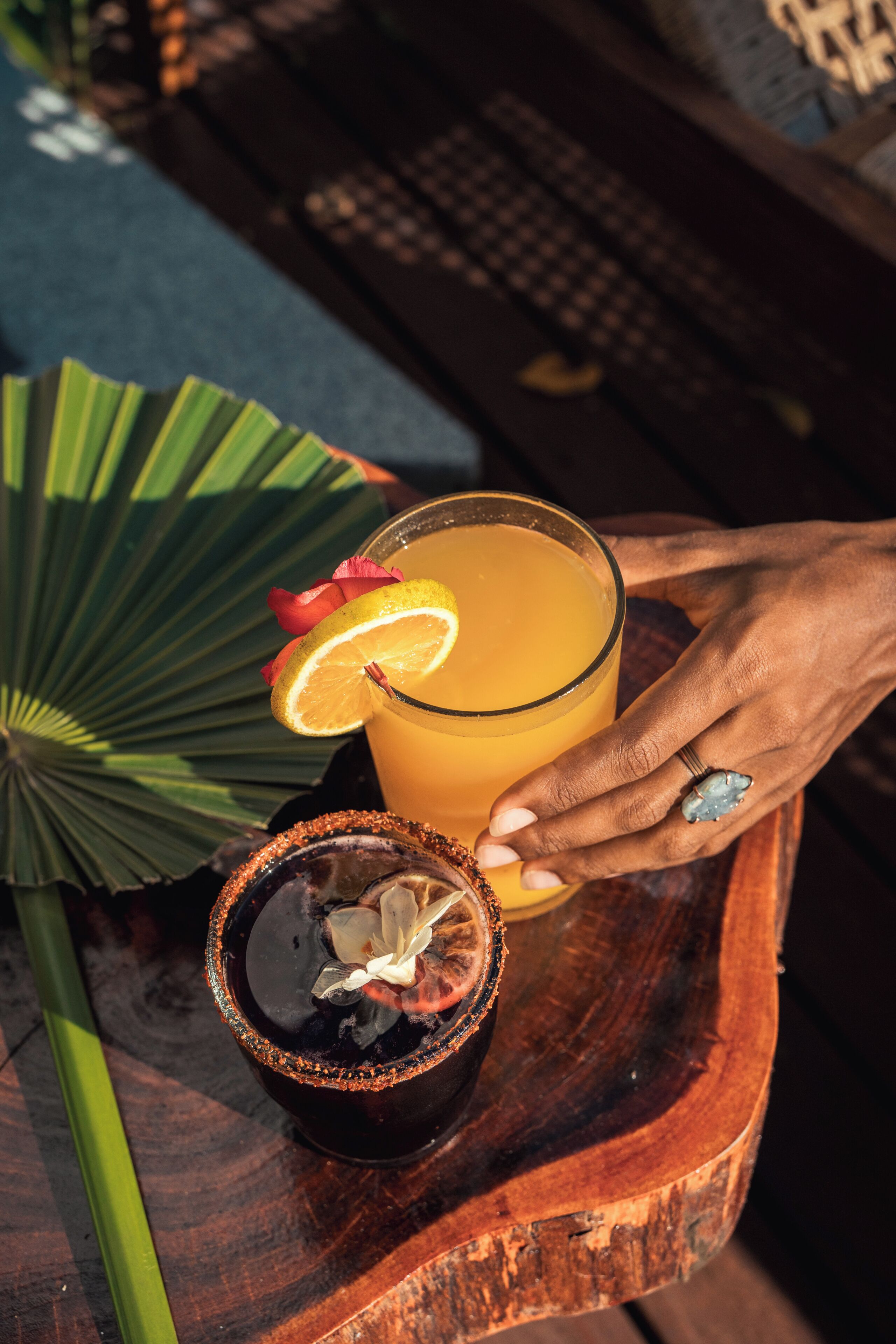A hand holding a glass of orange juice with a slice of lemon on a wooden tray.