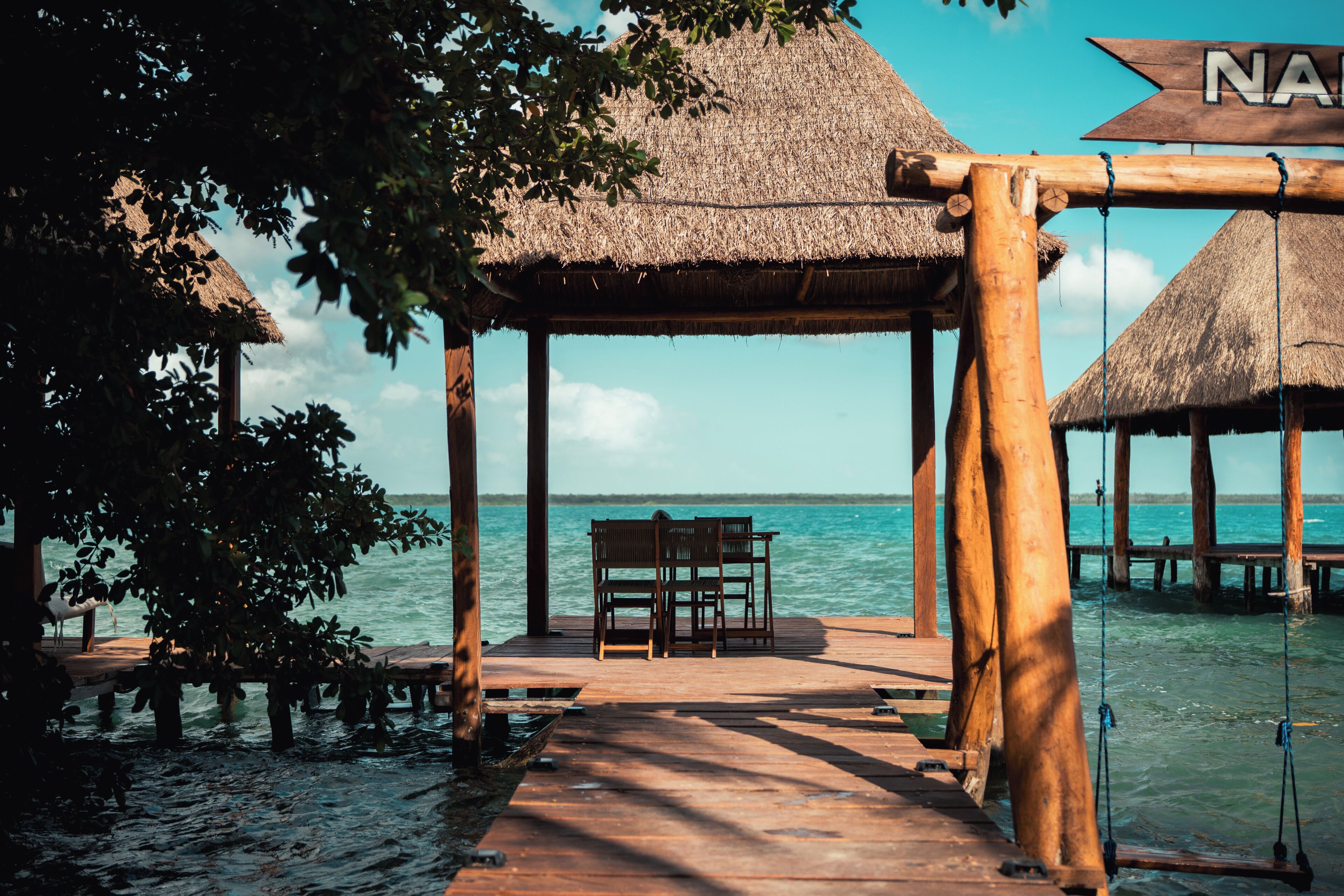 A wooden pier leading to a thatched-roof structure with a bench.