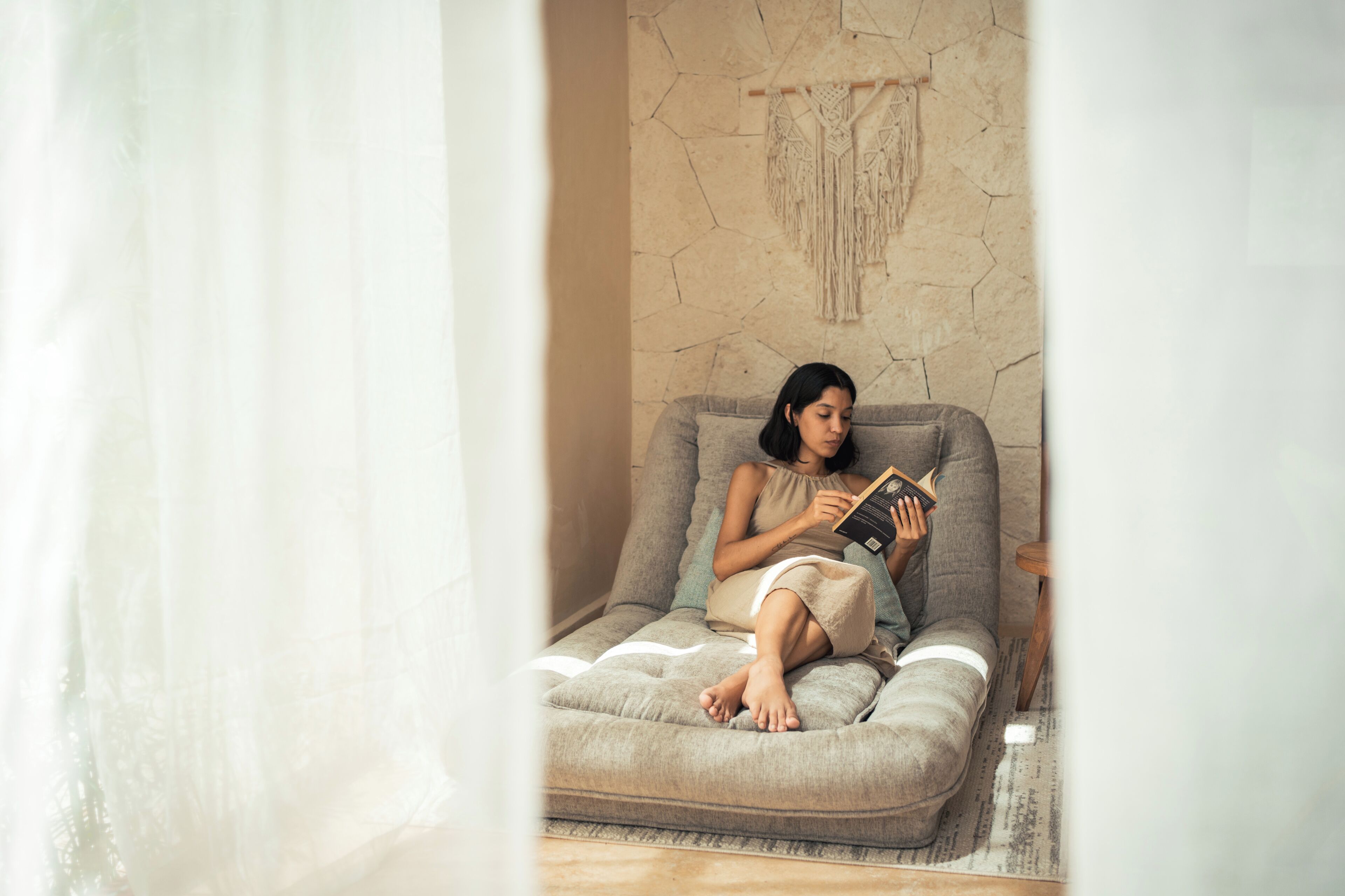 A person sitting on a sofa with a book, in a room with a textured wall and a curtain.
