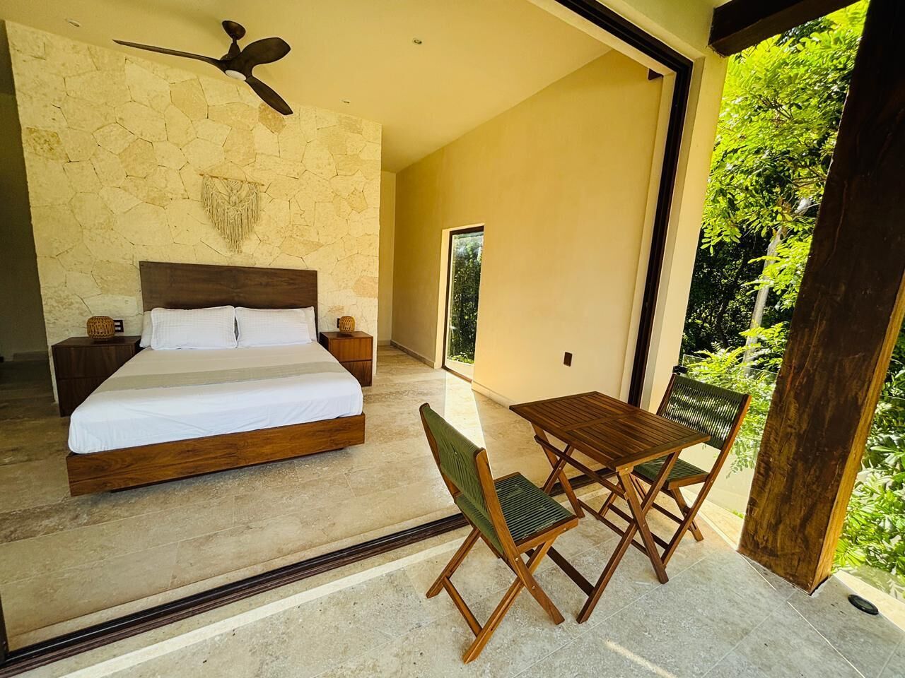 A bedroom with a bed, bedside tables, a ceiling fan, and a view of greenery outside.