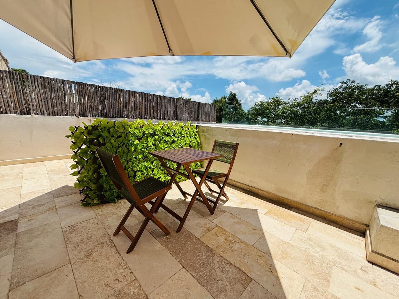 Patio esterno con tavolo e due sedie, un ombrellone grande e un siepe verde.