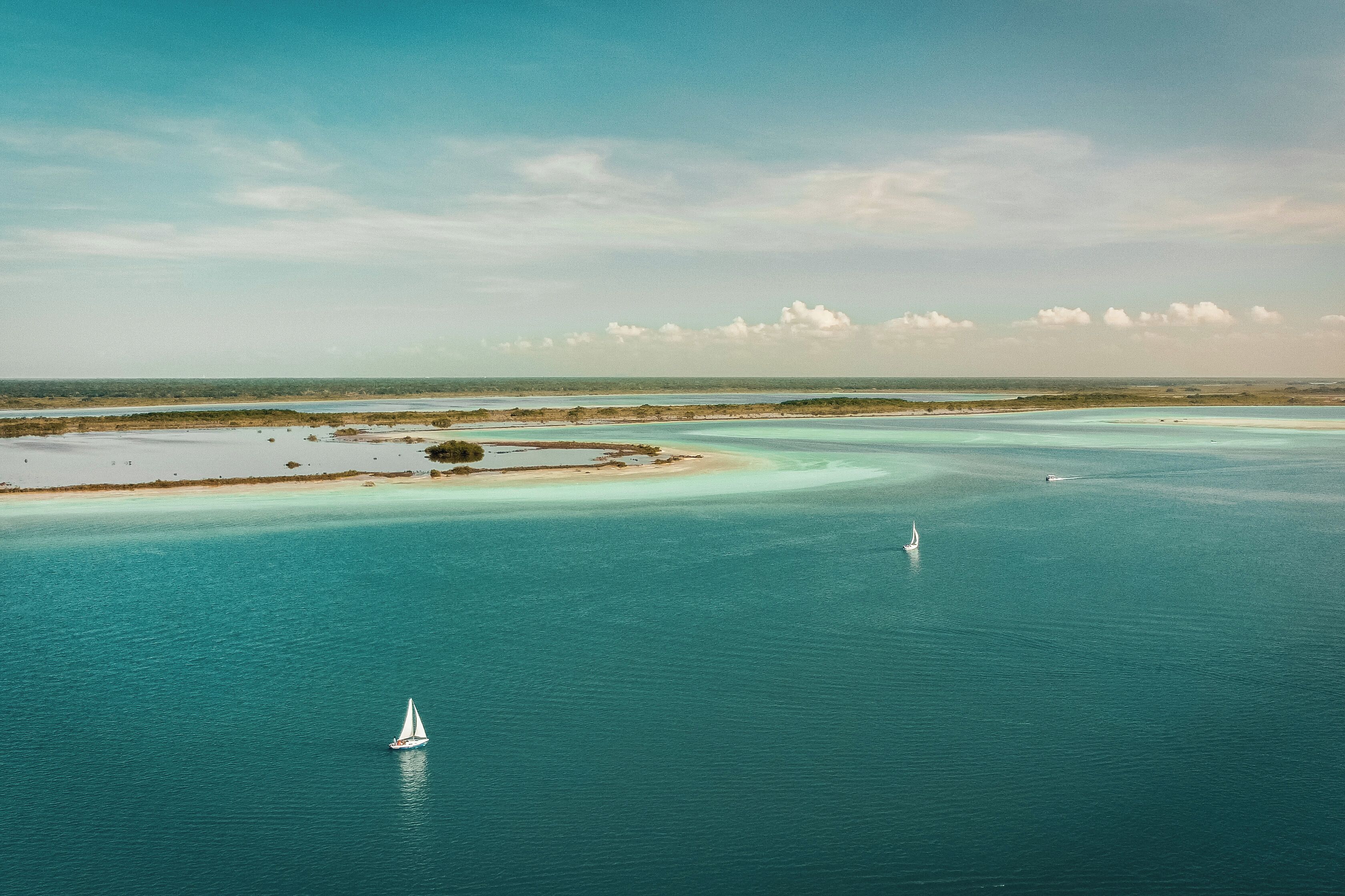 A coastal landscape with clear turquoise waters, a sandy beach, and a small sailboat.