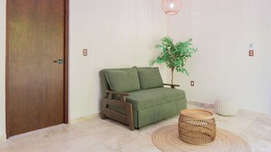Une pièce avec une porte en bois, un canapé vert, une petite table en bois, un tabouret en osier et un luminaire suspendu.