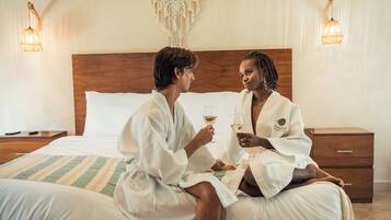Dos personas en una habitación de hotel, sentadas en una cama con cabecera de madera, sosteniendo copas de vino.