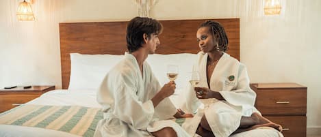 Two individuals in a hotel room, seated on a bed with a wooden headboard, holding wine glasses.