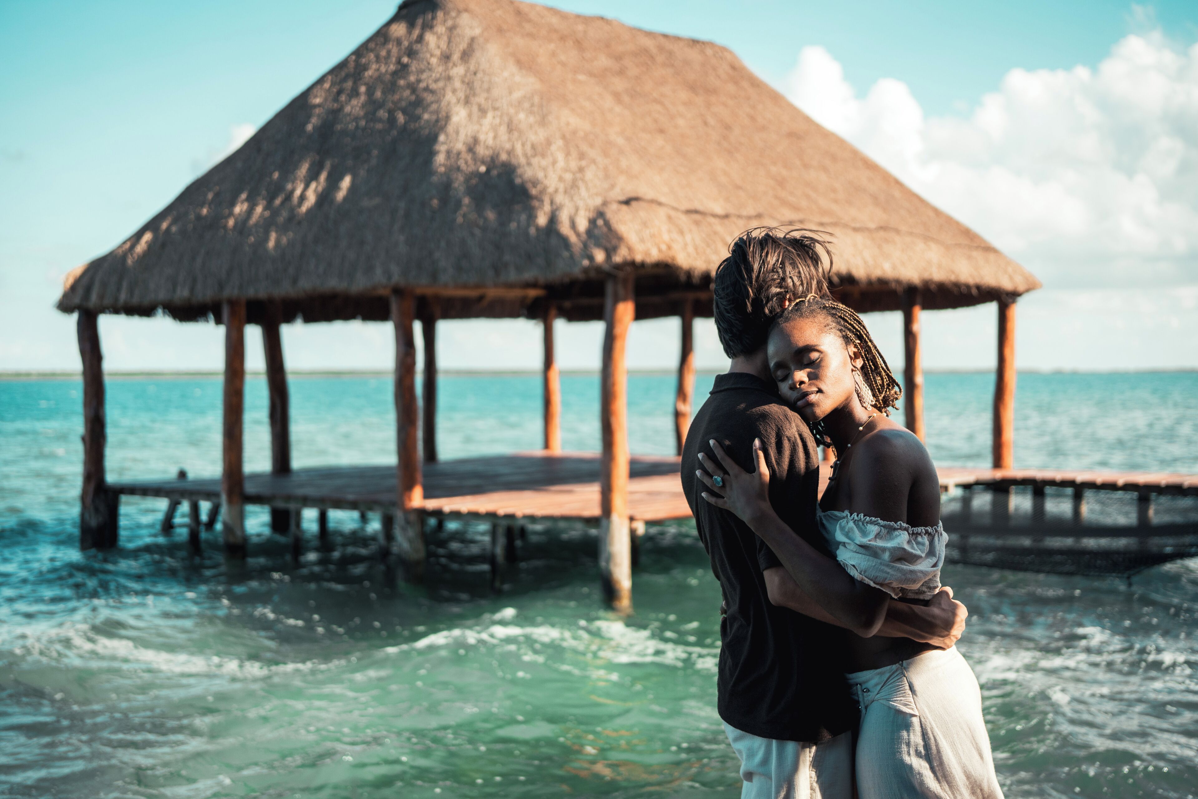 A couple embracing on a wooden pier with a thatched-roof structure in the background.