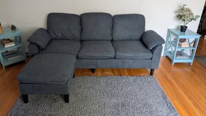 Ein graues Sofa mit passendem Hocker, ein türkisfarbener Beistelltisch mit Büchern und ein Blumentopf an einer weißen Wand.