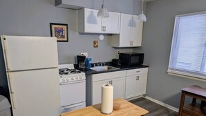 Uma cozinha compacta com armários brancos, fogão a gás, micro-ondas e uma pia. Há uma pequena mesa com um rolo de papel toalha e um banquinho de madeira.