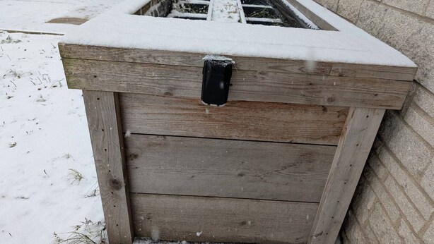 Estrutura de madeira com uma trava preta, parcialmente coberta por neve, em frente a uma parede de tijolos.