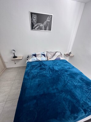 A bedroom with a bed, a nightstand, a wall-mounted picture, and a small shelf with a plant.