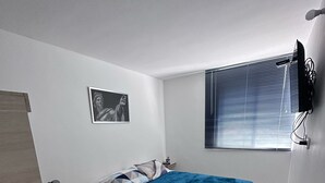 A bedroom with a bed, a bedside table, a lamp, a picture on the wall, and a window with blinds. - Apartment Departure for Salento and Filandia (Armenia)
