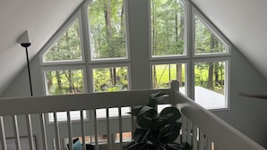 A room with a large window overlooking a forested area, a white railing, and a large plant in the foreground. - Cozy, family friendly, home away from home in welcoming Ocean Pines! (Ocean Pines)