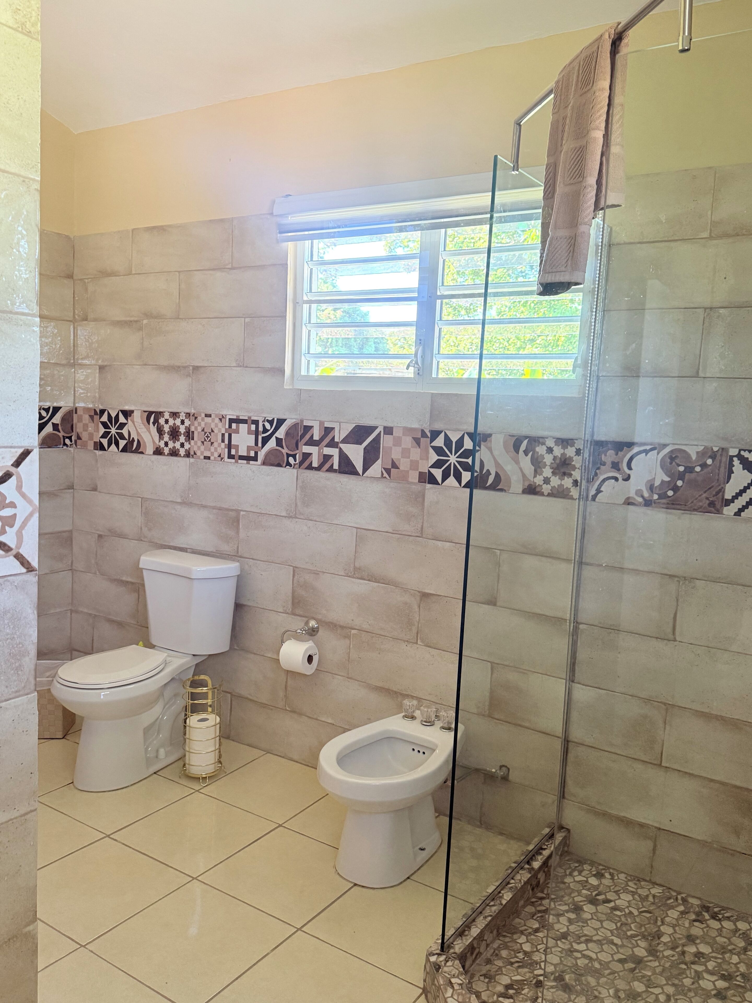 Un baño con dos inodoros, un bidé, una ducha con puerta de vidrio y azulejos estampados.