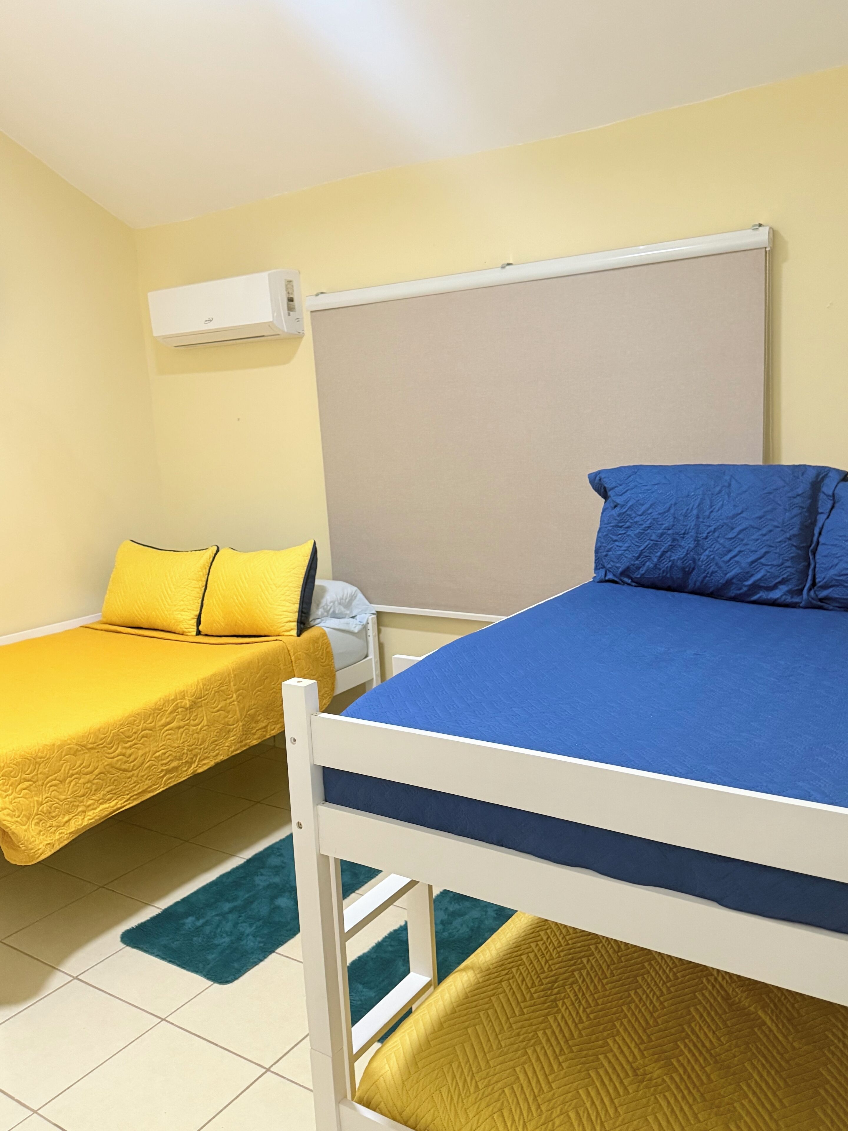 A room with two bunk beds, one with a blue mattress and the other with a yellow mattress. There is a white air conditioning unit on the wall above the beds.