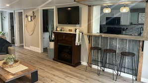 Una sala de estar moderna con chimenea, piso de madera y una cocina visible a través de una puerta abierta.