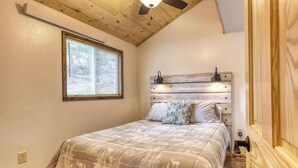 A bedroom with a wooden ceiling, a bed with a patterned bedspread, a window with blinds, and a nightstand with a lamp.