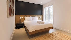 Un dormitorio moderno con una cama grande, cabecera de madera, mesita de noche pequeña y un ventanal que proporciona luz natural.