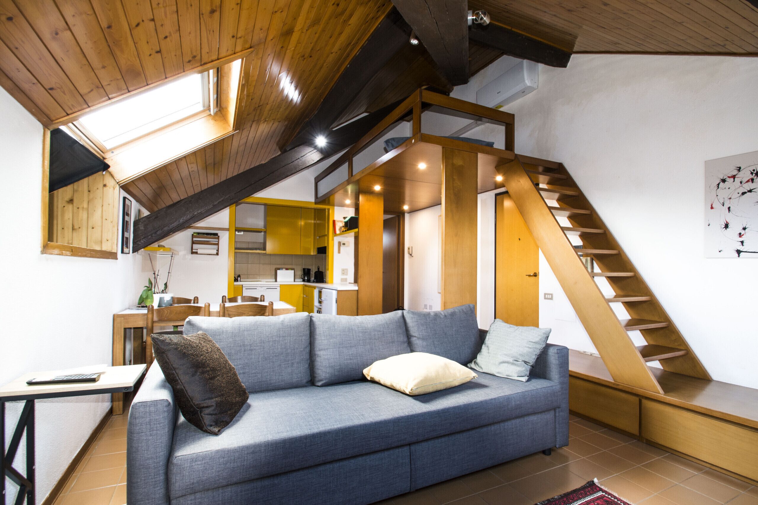 Een moderne woonkamer met een blauwe bank, een houten trap en een keuken op de achtergrond.