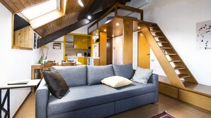 A modern living room with a blue sofa, a wooden staircase, and a kitchen in the background.