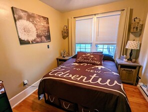 A bedroom with a bed featuring a 'Adventures to New' themed bedspread, a window with blinds, a bedside table with a lamp, and a wall-mounted light fixture.