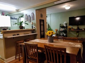 A dining area with a wooden table and chairs, a kitchen area with a 'Kitchen' sign, and a television mounted on the wall.