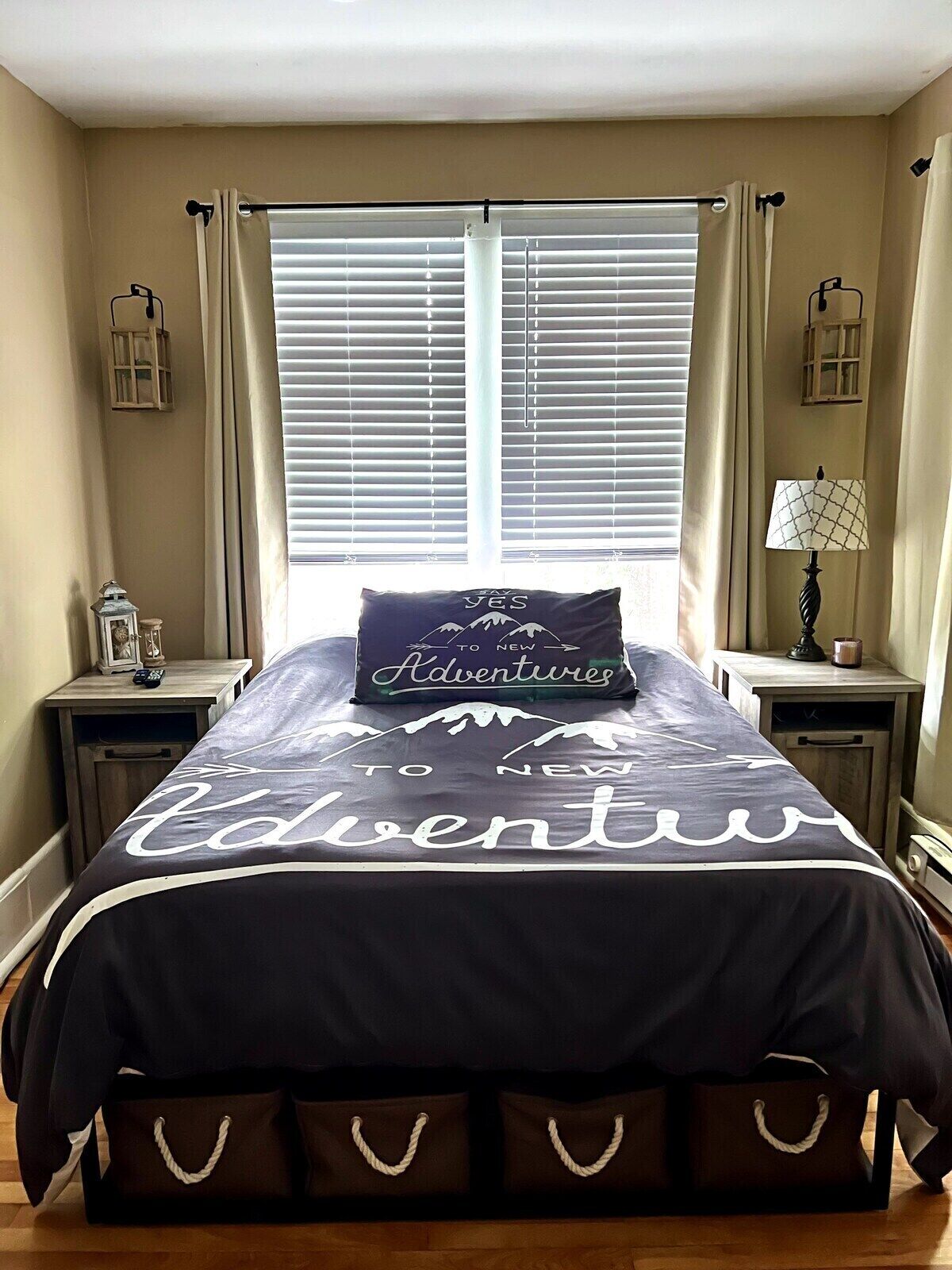 Une chambre à coucher avec un lit agrémenté d’une couvre-lit sur le thème « À l’aventure ! », une fenêtre avec des stores et une table de chevet sur laquelle se trouvent une lampe et une lanterne.