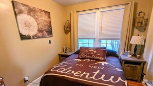A bedroom with a bed, a window with blinds, a nightstand, a lamp, and a wall decoration.