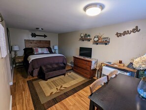 A bedroom with a bed, dresser, desk, and TV. There is a wall clock, a shelf with decorative items, and a sign that says "travel". - Robin's Nest Charming Whole House Rental (Saranac Lake)
