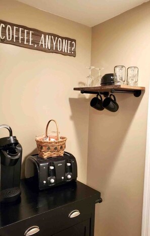 Un coin cuisine équipé d’une machine à café, d’un grille-pain et d’un panier posé sur le comptoir. Une étagère au-dessus contient des verres, et une pancarte indique : « CAFÉ, POUR QUI ? ».