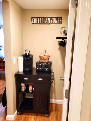 A kitchenette with a coffee maker, toaster, and basket on a cart, and a sign above that says "COFFEE, ANYONE?".