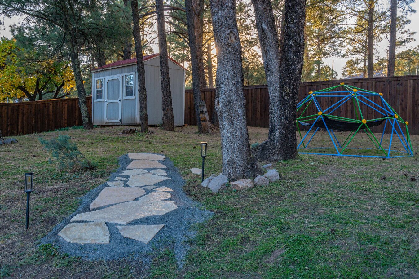 A stone pathway leading to a small shed, surrounded by trees and a wooden fence.