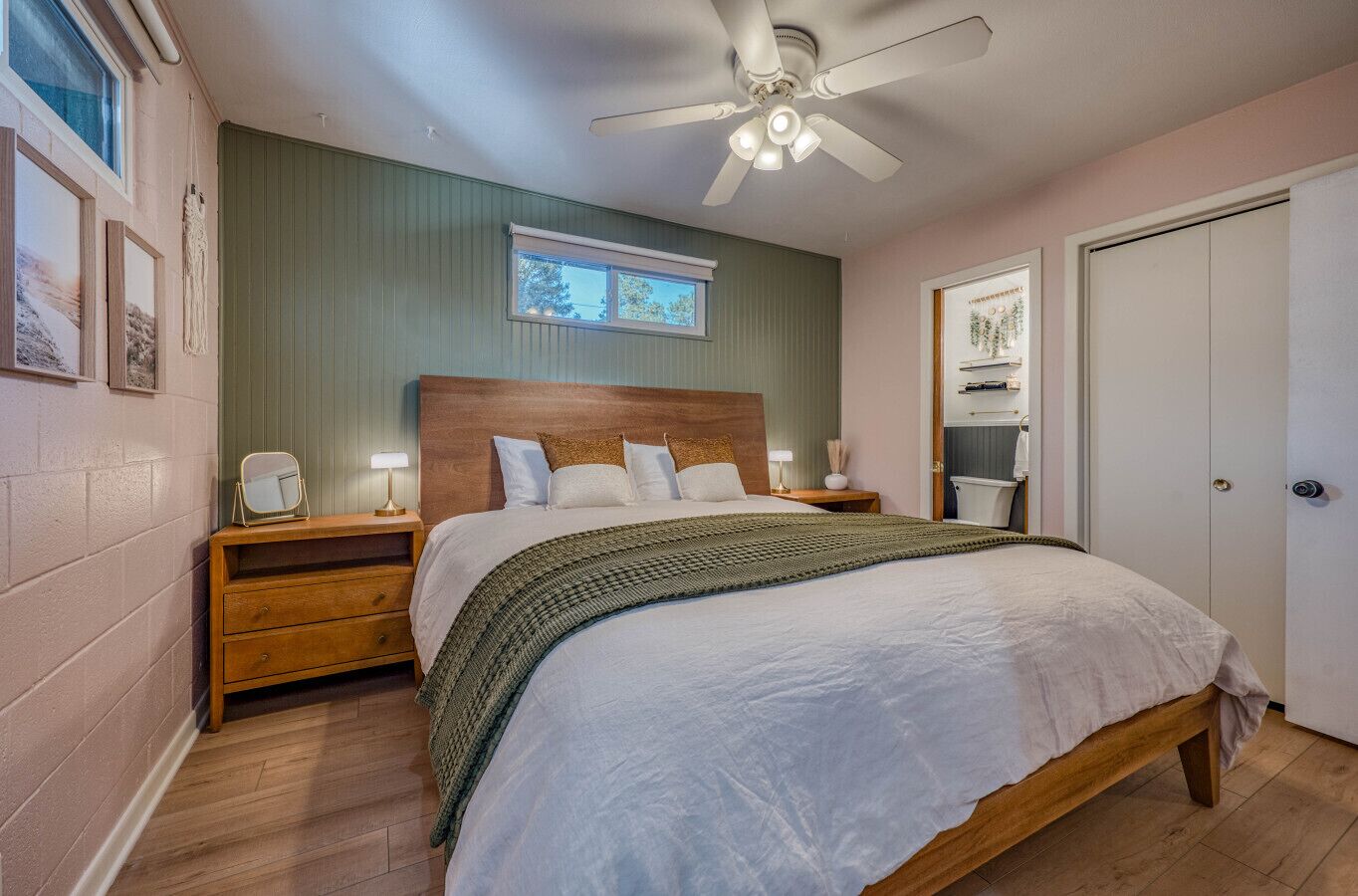 A bedroom with a wooden bed, a nightstand with a lamp, a ceiling fan, and a window with a view of trees.
