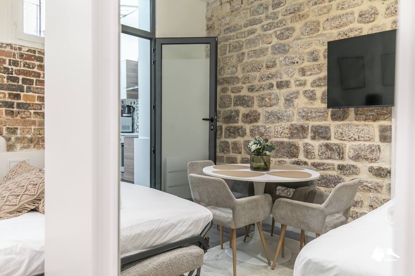 Une pièce de taille modeste avec un mur en briques, une table ronde entourée de chaises, un lit et un téléviseur fixé au mur.