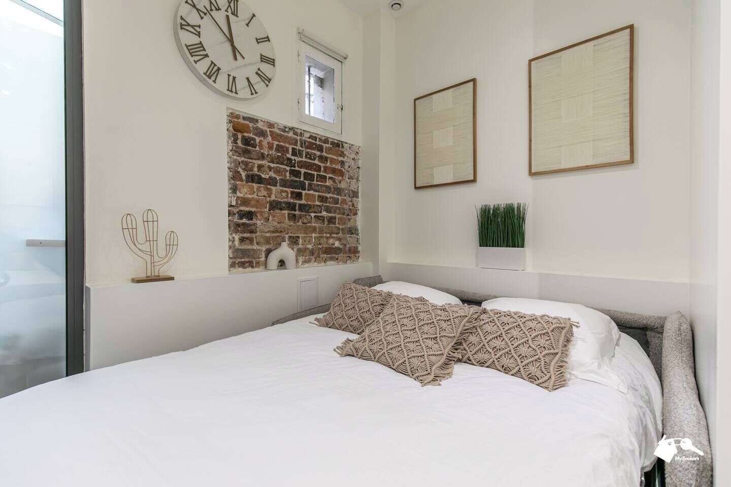 A bedroom with a brick wall, a large wall clock, two framed pictures, and a cactus decoration.