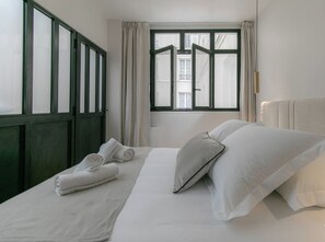 Ein ordentlich bezogenes Bett mit weißen Leinen und Kissen, ein Fenster mit Vorhängen und Blick ins Freie.