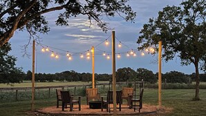 An outdoor seating area with wooden chairs and tables, surrounded by a string of lights, set against a backdrop of trees and a grassy field. - Windy Hill Farmhouse in Caldwell (Caldwell)