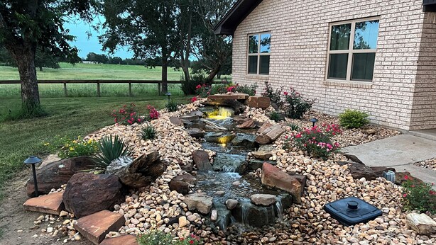 A landscaped garden with a water feature, rocks, and flowers in front of a brick building. - Windy Hill Farmhouse in Caldwell (Caldwell)