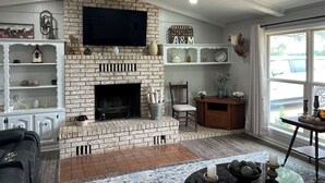 A living room with a brick fireplace, a flat-screen TV mounted above it, and a large window providing a view of the outside. - Windy Hill Farmhouse in Caldwell (Caldwell)