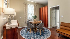Een eetruimte met een ronde tafel, vier stoelen, een blauw tapijt met patroon, een houten buffetkast met een lamp, en een deur naar een andere kamer.