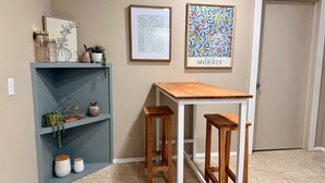 Een keuken met een houten tafel en twee krukken, een blauwe plank met decoratieve items, en twee ingelijste kunstwerken aan de muur.