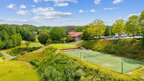 Ein Tennisplatz, umgeben von üppigem Grün, ein Parkplatz und ein Gebäude mit rotem Dach.
