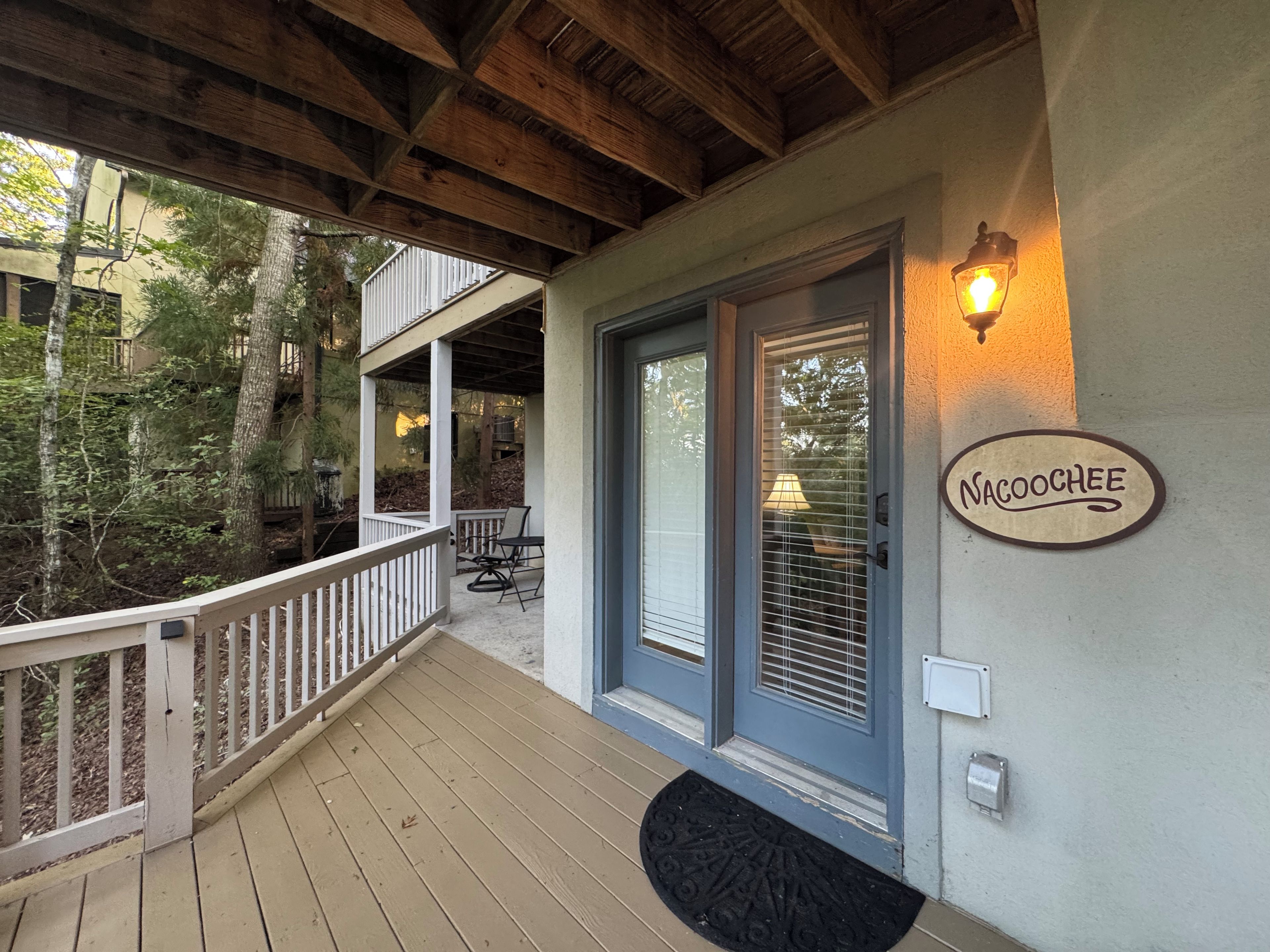 Une terrasse en bois avec une balustrade blanche, une porte dotée d’une fenêtre et une enseigne fixée au mur sur laquelle on peut lire « NACOOCHEE ».