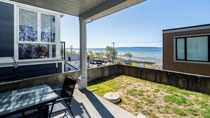 En overdækket terrasse med et bord og stole, udsigt til stranden, og en bygning med store vinduer.
