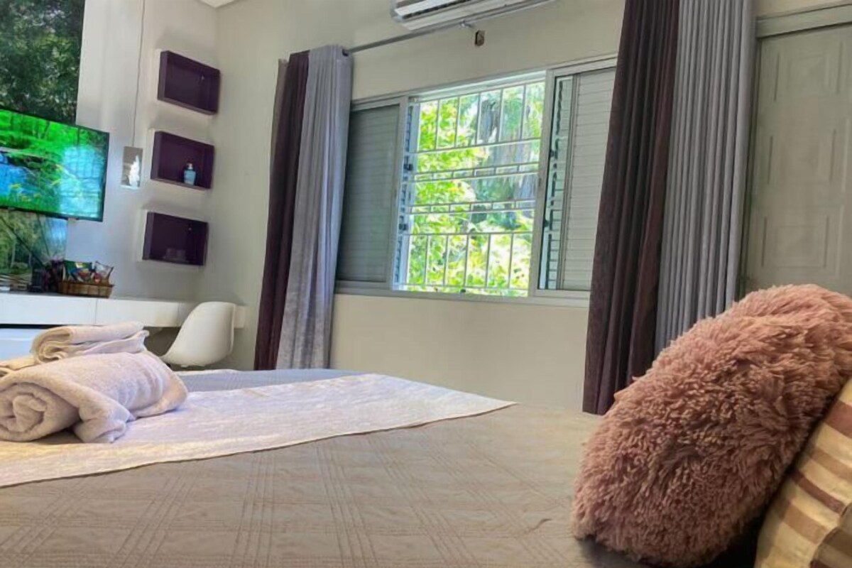 A hotel room with a bed, a TV, a window with curtains, and a wall-mounted shelf with decorative items.