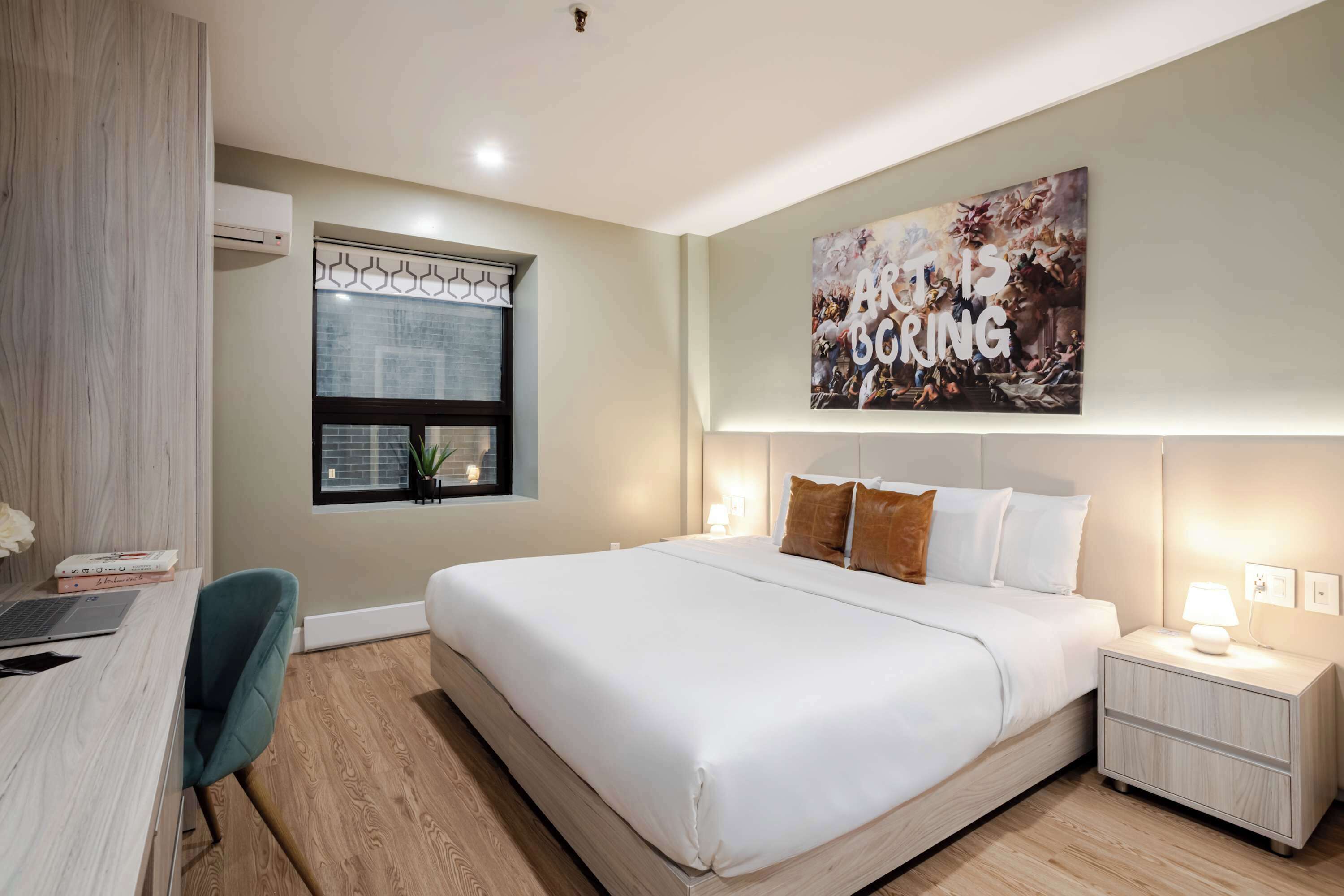 Une chambre d’hôtel moderne dotée d’un grand lit, d’un bureau avec un ordinateur portable, d’une fenêtre avec stores, d’un climatiseur fixé au mur et d’une affiche inspirante.