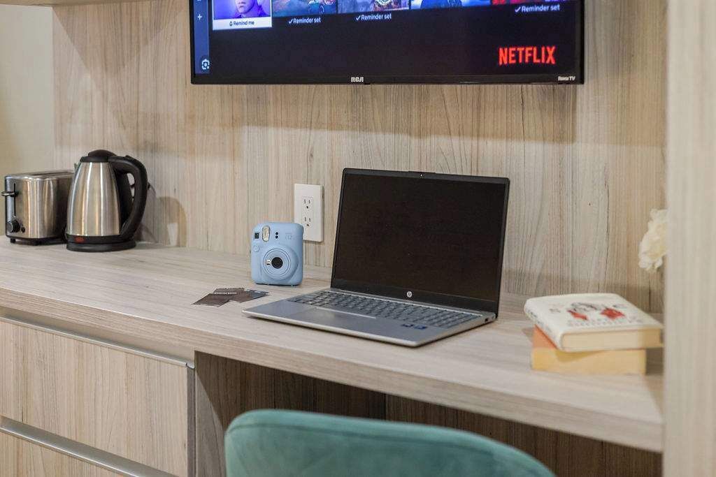 Habitación de hotel con escritorio de madera, una computadora portátil, una cámara, hervidor de agua y estantería.